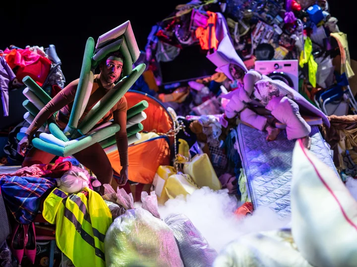Zwei Performer:innen in Kostümen aus Matratzen und Schaumstoff stehen zwischen aufgeschichteten Kleidungsbergen und Alltagsobjekten auf der Bühne.