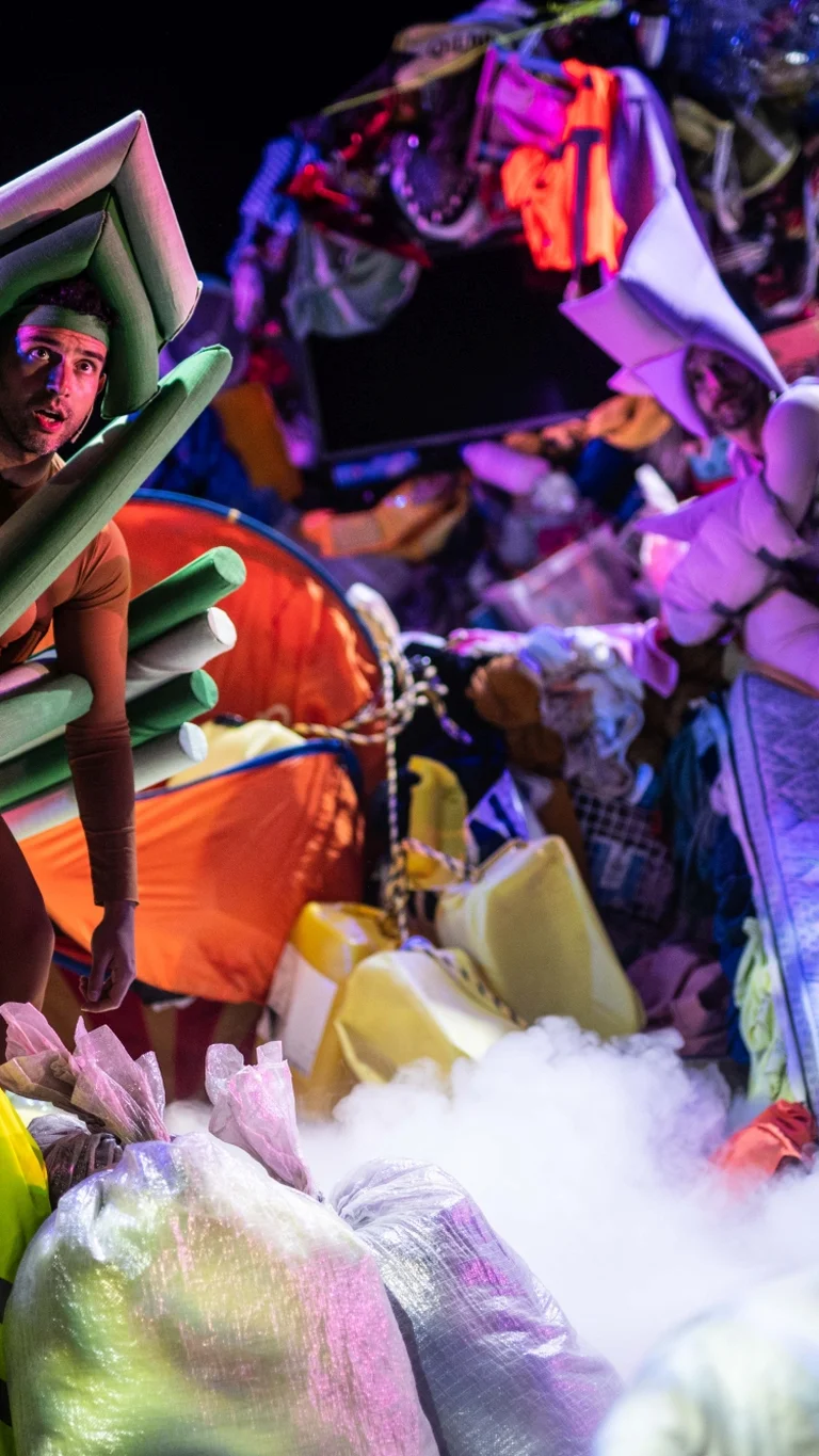 Zwei Performer:innen in Kostümen aus Matratzen und Schaumstoff stehen zwischen aufgeschichteten Kleidungsbergen und Alltagsobjekten auf der Bühne.