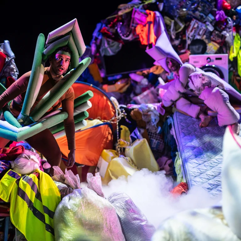 Zwei Performer:innen in Kostümen aus Matratzen und Schaumstoff stehen zwischen aufgeschichteten Kleidungsbergen und Alltagsobjekten auf der Bühne.