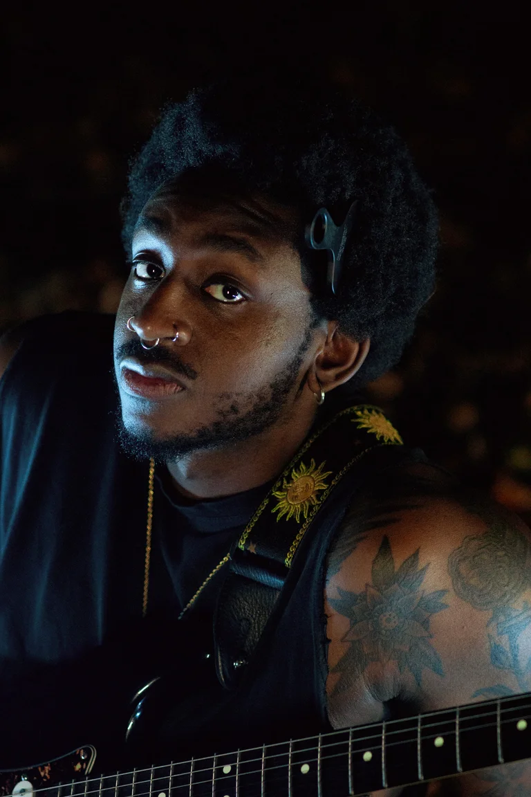 The musician Joël-João is sitting in a dark environment. He is wearing a black sleeveless shirt, a gold necklace, and has multiple facial piercings. A guitar strap with sun motifs is visible, as well as a black electric guitar in the lower part of the image.