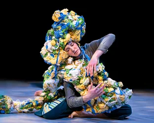 Eine Performerin sitzt auf dem Boden und ist mit einer großen Skulptur aus zerknülltem, mehrfarbigem Material bedeckt, die ihren Kopf und Körper umhüllt.