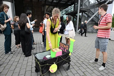 A woman in a yellow dress interacts with a colorful cart labeled "Teens Talk." Young people stand around her, chatting.