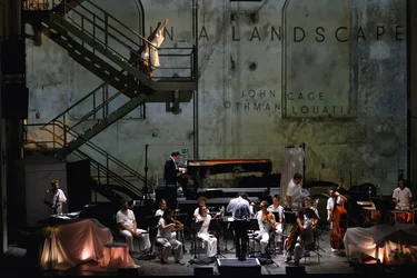 In a large industrial hall, the ensemble Les Apaches ! performs on stage. The musicians are dressed predominantly in white and play instruments including piano, strings, wind instruments and percussion. On the left, a large metal staircase runs along the wall, with a person dressed in white moving on it. The words “IN A LANDSCAPE” appear in large letters on the concrete wall.