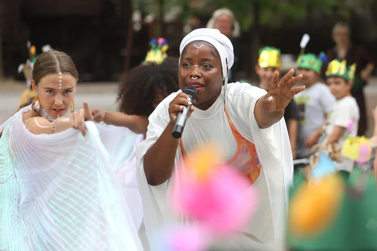 A musician sings, she holds her hand in the air and looks into the audience. A dancer dances in white, looking directly into the camera