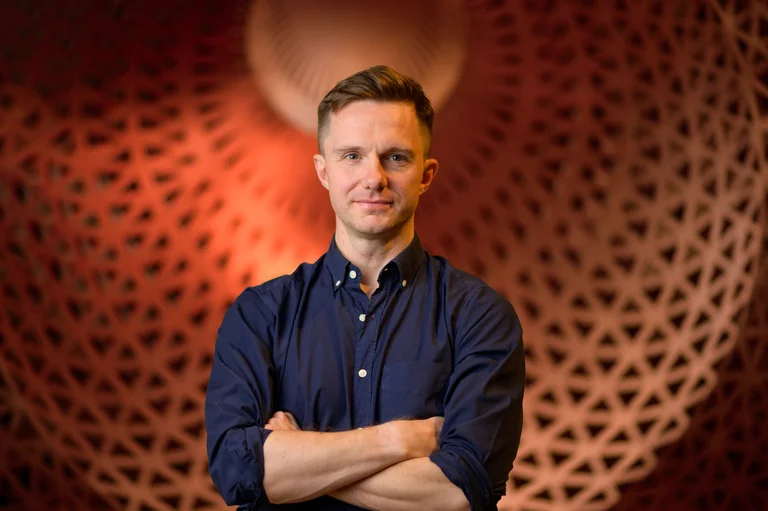 James McVinnie stands with his arms crossed in front of an artistically patterned, orange-brown lit background. He is wearing a dark blue shirt with rolled-up sleeves and has a confident, friendly expression.