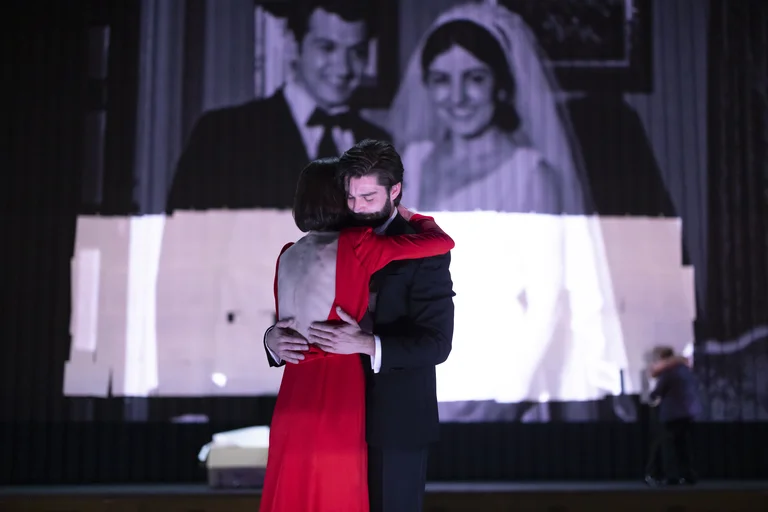 Eine Frau in einem roten Kleid mit freiem Rücken und ein Mann im Anzug umarmen sich. Im Hintergrund wird ein Schwarzweißfoto eines lächelnden Paares, vermutlich an ihrem Hochzeitstag, projiziert