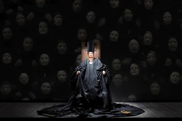 Scene from Balkan Erotic Epic: A figure draped in black sits on a throne in front of a wall of projected faces. The atmosphere is solemn and dark.