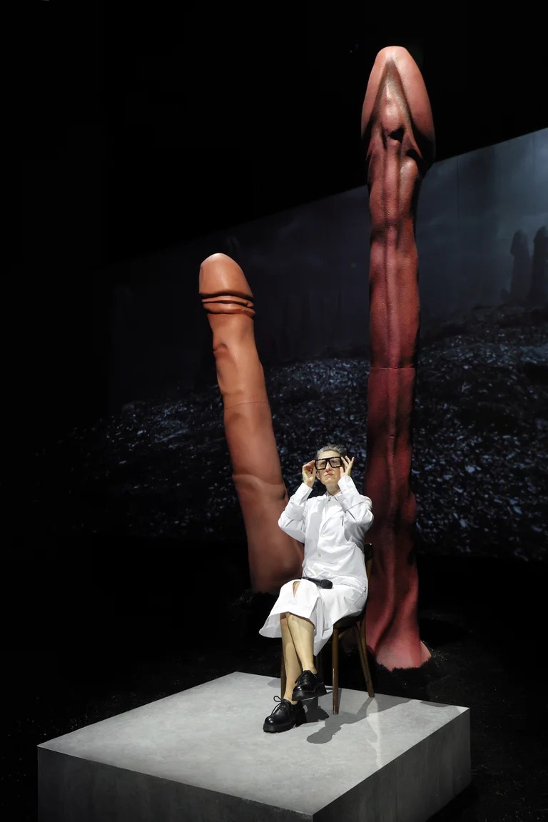 Woman in a white coat sits on a platform in front of two oversized, phallic sculptures in a dark setting.