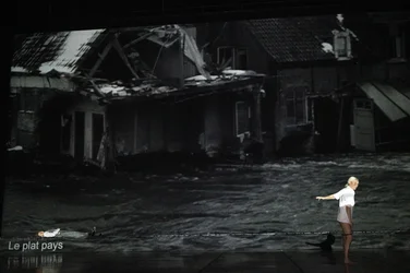 Anne Teresa De Keersmaeker steht auf der Bühne vor einer großflächigen Projektion eines überfluteten Gebäudes; links liegt Solal Mariotte am Boden.