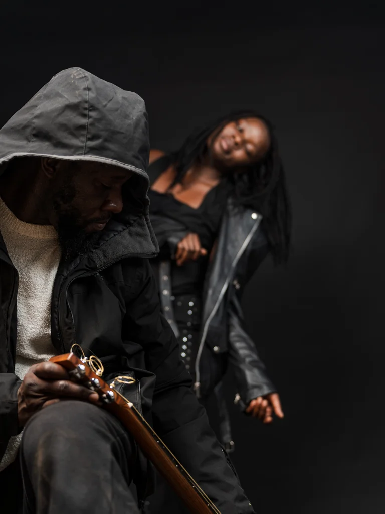Jean-Robert Koudogbo Kiki sits in the foreground with his gaze lowered, holding a guitar in his hands, dressed in a hooded jacket. In the blurred background, Niaka Sacko leans slightly backward in black clothing, her head tilted to the side.