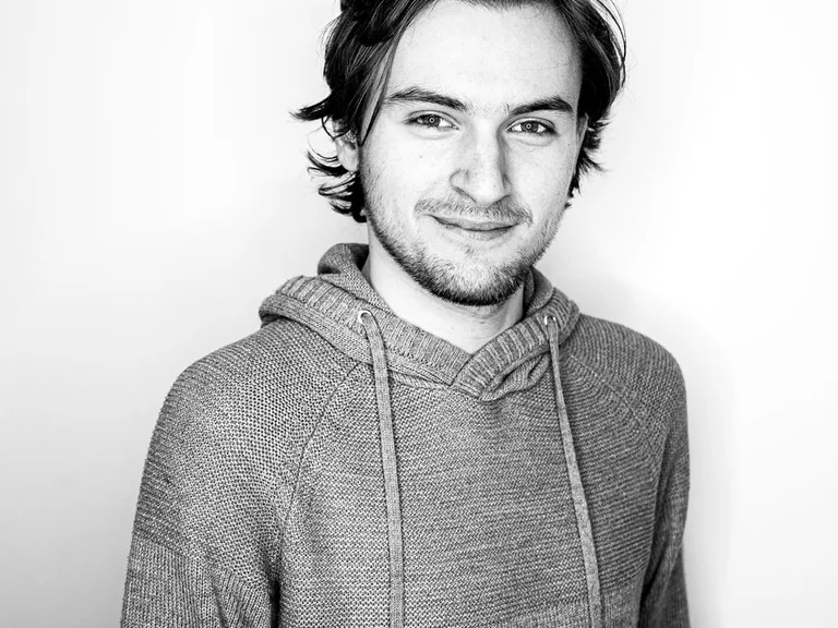 Dominic Bade mit mittellangen Haaren und leichtem Bart in Kapuzenpullover vor hellem Hintergrund.