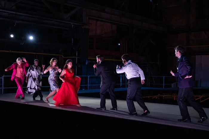 Several performers move dynamically across a brightly lit, elevated stage inside a darkened industrial hall. On the left side of the image, Helena Rasker wears a vibrant pink suit, Claron McFadden appears in a black-and-white patterned robe, Nina van Essen sparkles in a silver sequin dress, and Sophia Burgos stands out in a flowing red evening gown. Opposite them, male performers dressed in elegant evening attire engage with energetic gestures, creating a sense of theatrical tension and movement.