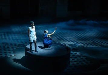 Two performers are on a circular platform in a dark, blue-lit stage scene surrounded by mist. One stands in white clothing and black boots, while the other, dressed in blue, kneels and raises one arm.