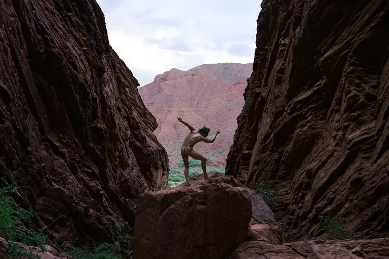 A canyon with steep, dark rock walls that open up to reveal a reddish mountain landscape in the background. In the center of the image, a naked person stands on a large rock, striking an expressive dance pose with outstretched arms and a bent leg.