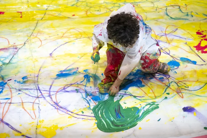 A child kneels on a painted floor and spreads green paint onto the canvas with their hands. The surface is already covered with yellow, blue, and red marks.
