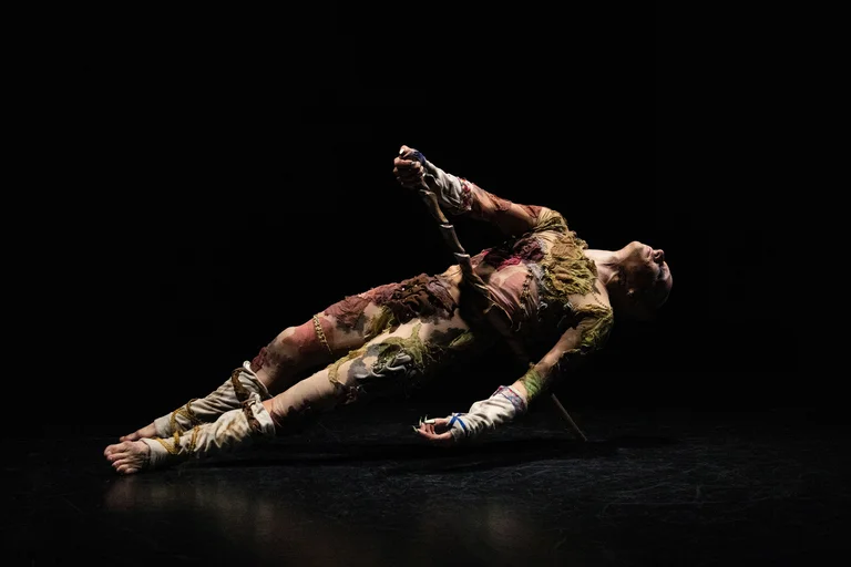 A dancer in a colorful, tattered costume lies diagonally on their back, supported by a stick. The scene is dark, with the body dramatically lit.