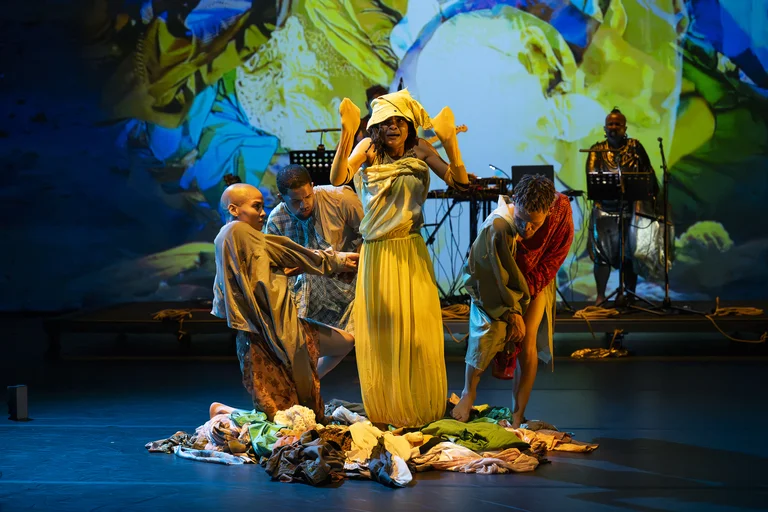 Several performers surround a person in a yellow skirt standing at the center of the stage on a pile of clothes, with colorful projections in the background.