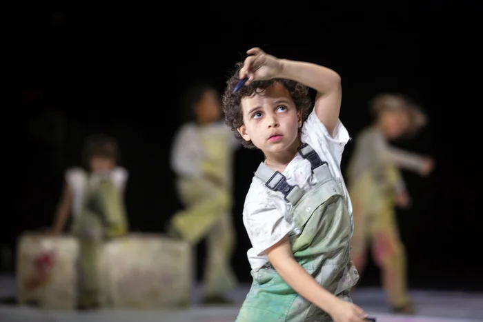 A child with curly hair and a white overall with paint stains raises one arm with a brush in the air. In the background, other children in similar outfits are visible on stage.