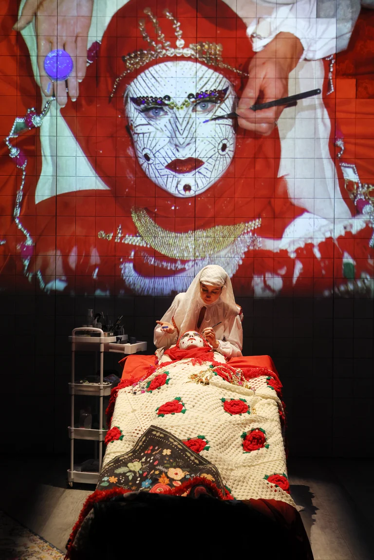 Tiled stage room: one person lies under a colorful embroidered blanket as another leans over applying makeup; above them, a giant projection of a red-veiled face.