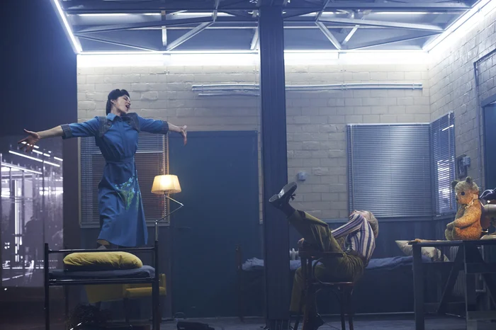 Stage scene: An actress stands with outstretched arms on a bed, another actress leans back on a chair; a teddy bear sits on the table.