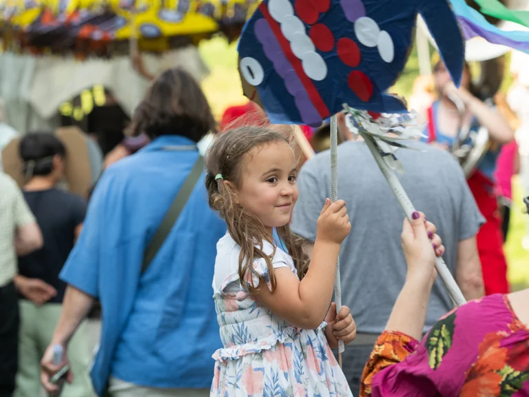 A joyful girl with long brown hair holds a colorful, handmade paper fish on a stick. She wears a summery floral dress and smiles while looking to the side. In the background, many people dressed in colorful clothing participate in a festive parade or procession.