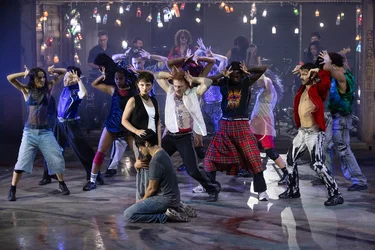Two performers stand close together on a stage. The person in the foreground is kneeling; the person behind has placed their hand on the head of the person in front. In the background, performers dance in front of a shipping container where a band is playing beneath strings of lights.