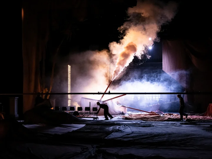 Bühne einer Performance im Dunkeln, auf der dichter Rauch aus einer schräg aufragenden Konstruktion strömt. Zwei silhouettenhafte Personen bewegen sich über große Stoffbahnen im Vordergrund, während starke Lichtquellen den Rauch dramatisch beleuchten.