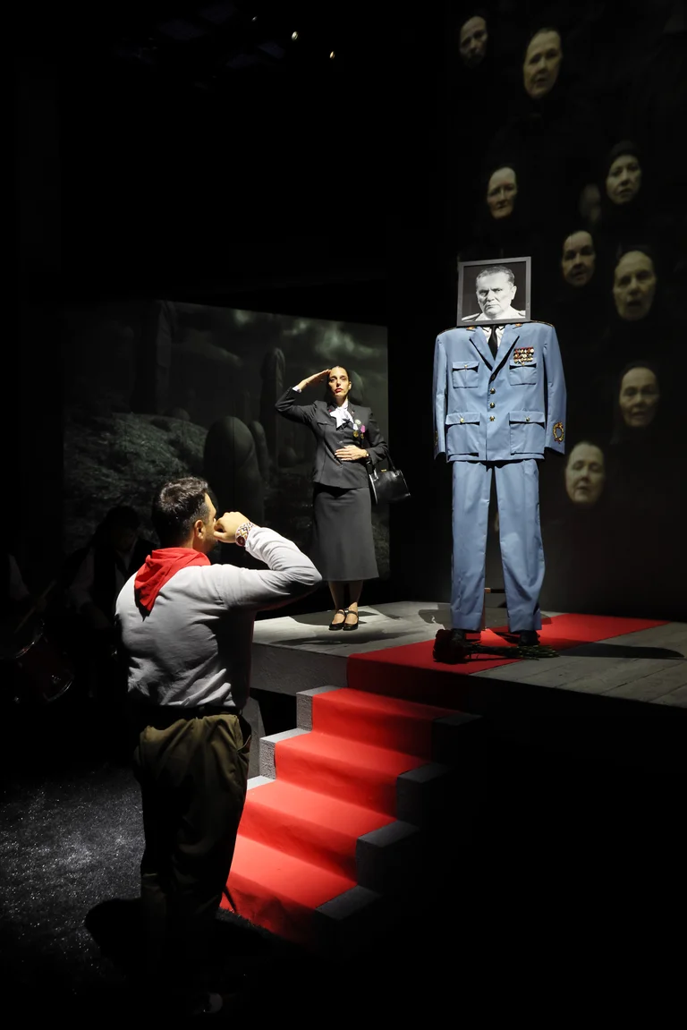 Dark stage scene with salutes, red steps, a blue-uniform figure topped by a portrait, and floating faces in the background.