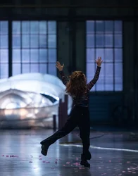 A person stands on a stage with their arms raised and one leg lifted. Pink flower petals are scattered across the floor. Large windows and an illuminated inflatable structure are visible in the background.