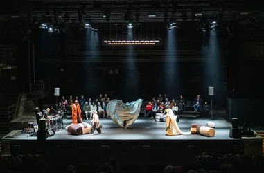 Several performers move across a large stage among oversized padded objects and a swirling fabric element. Musicians play at the side of the stage while audience members sit behind on the stage.