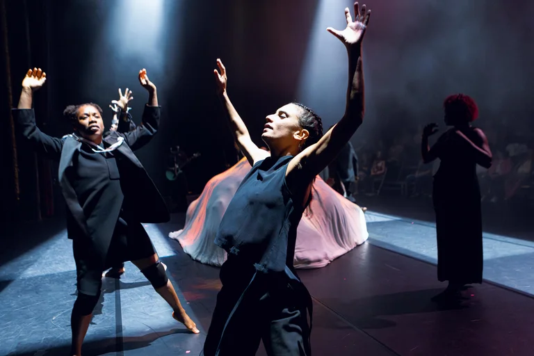 Two dancers stretch their arms into the air on stage. In the background of the dark stage lies a large, bright structure.