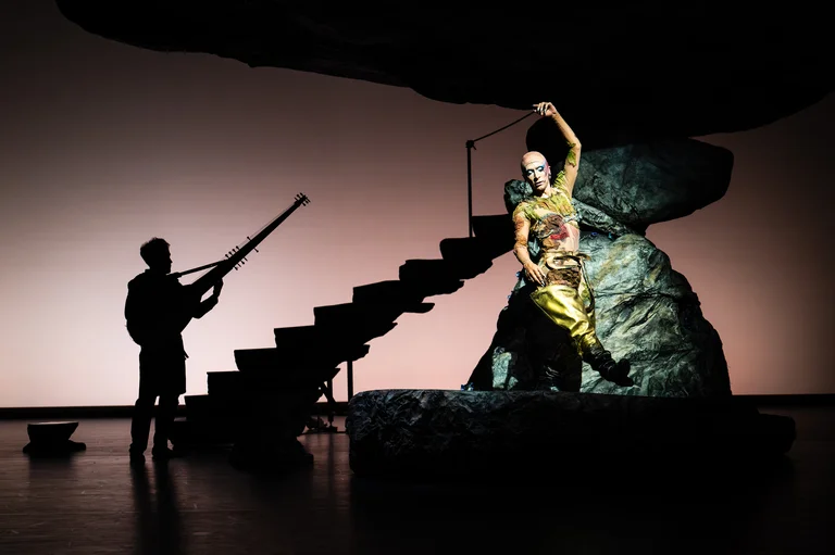 A performer stands on a rock, dressed in a golden costume with intricate details. On the left side of the image is the silhouette of a musician with a string instrument.