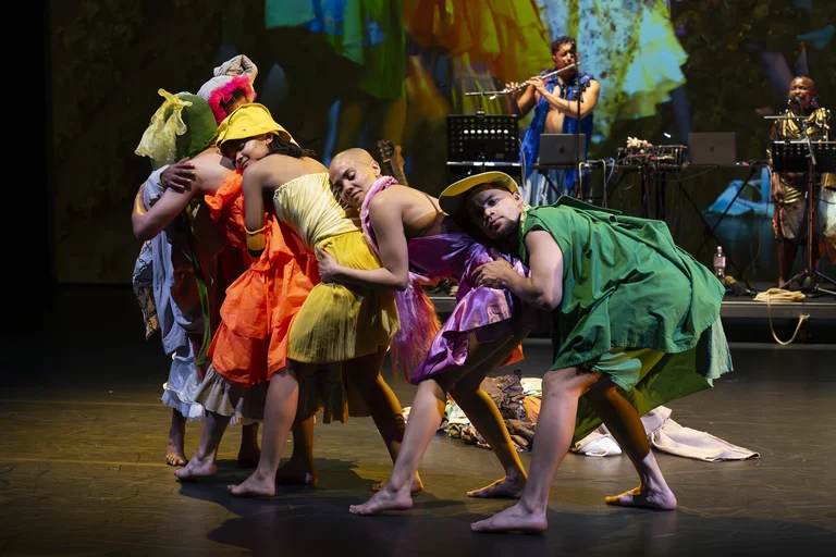 A group of performers in colorful dresses and costumes lean closely together on stage, with musicians playing in the background.