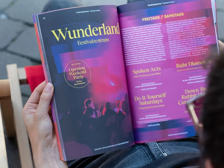 Eine Person hält ein aufgeklapptes Festivalprogramm der Ruhrtriennale, das farbig gestaltete Seiten mit Informationen zum Festivalzentrum Wunderland zeigt.
