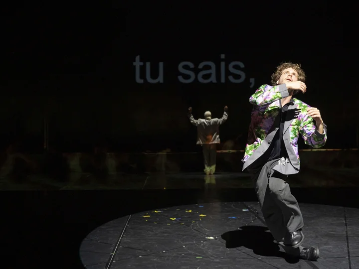Solal Mariotte in a grey suit moves on a circular, lit stage area; Anne Teresa De Keersmaeker stands in the background in front of projected text.