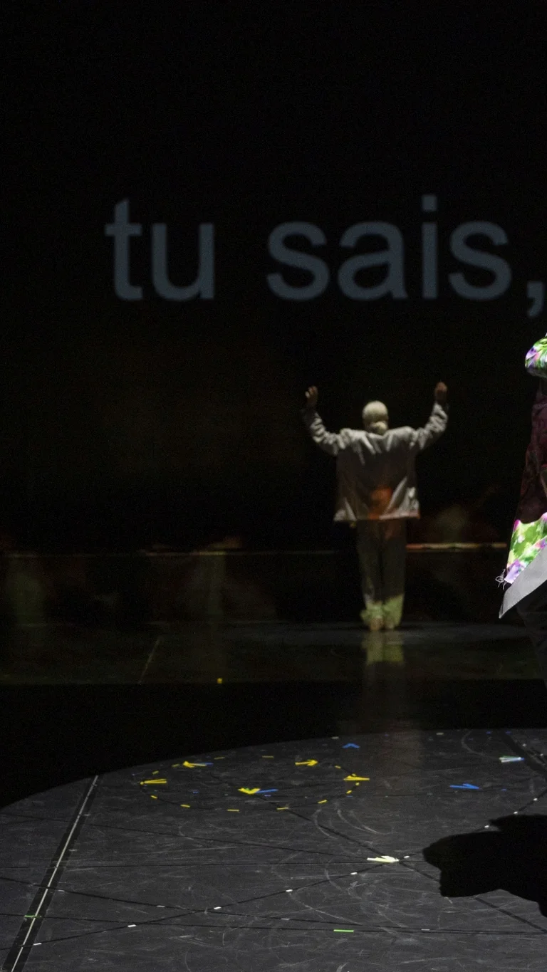 Solal Mariotte bewegt sich auf einer runden, beleuchteten Spielfläche in grauem Anzug; im Hintergrund steht Anne Teresa De Keersmaeker vor projiziertem Text.