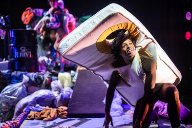 Performer mit einer Matratze als Kostüm kniet auf der Bühne zwischen Säcken, Textilien und Haushaltsgegenständen.