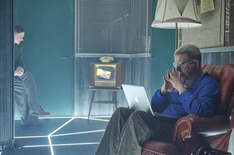 An actor sits in an armchair with a laptop, an old television in the background; another person leans against the door