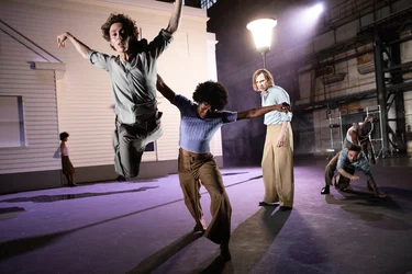 Several performers dance energetically on an open stage in front of a white house structure; one leaps dynamically into the air.