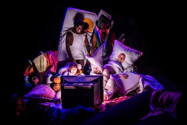 Mehrere Performer:innen sitzen und liegen auf einem Berg aus Decken, Kissen und Stoffen und schauen gemeinsam auf einen Monitor.