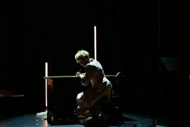Joyce DiDonato sits at a wooden table in a white dress on a dark stage, writing on a sheet of paper. Vertical light bars illuminate the otherwise dark scene.