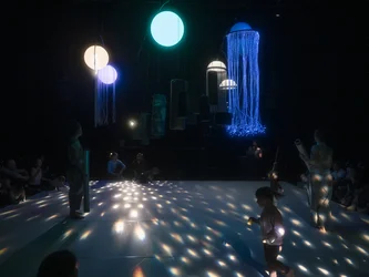 Kleinkinder und Erwachsene befinden sich auf einer dunklen Bühne unter großen leuchtenden Kugeln und Objekten mit langen Lichtfäden. Lichtpunkte spiegeln sich auf dem weißen Boden.