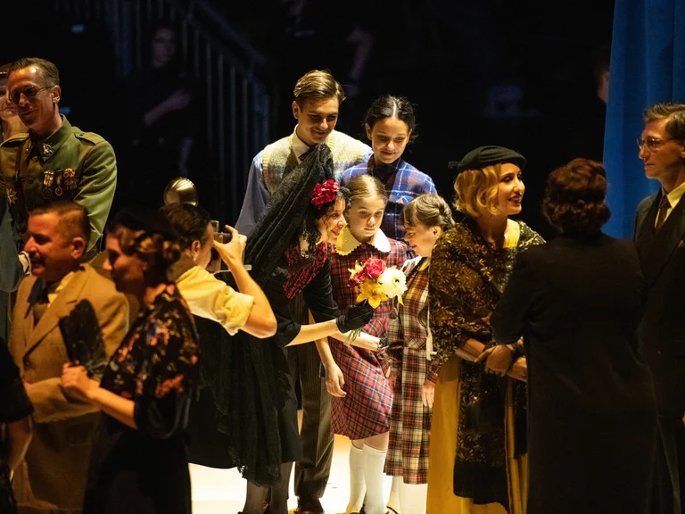 Eine Gruppe von Menschen in historischen Kostümen steht dicht beisammen, darunter Soldaten, Frauen und Kinder. Ein Mädchen im karierten Kleid hält einen Blumenstrauß, während andere Figuren lebhaft miteinander sprechen.