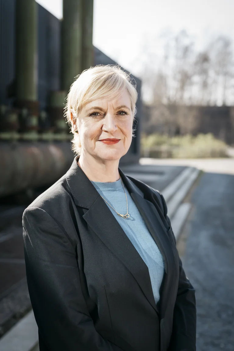 Anita van Dolen, curator/advisor dance, stands in front of an industrial backdrop and looks into the camera.