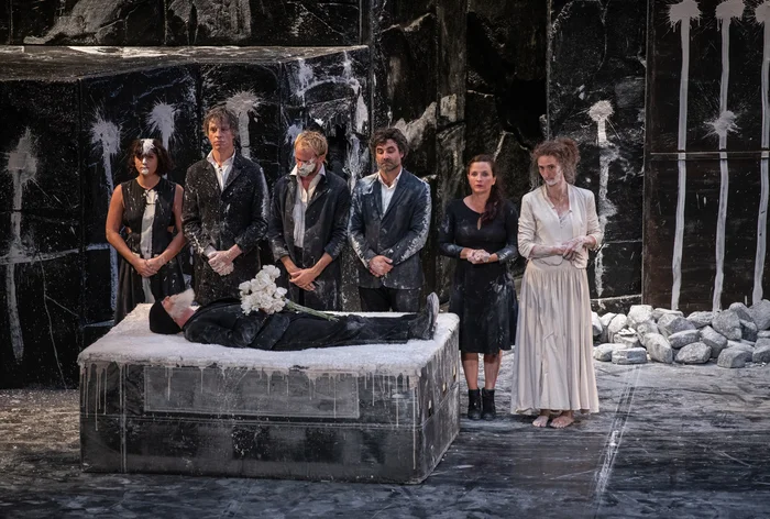 Six figures in black and white stand solemnly by a dust-covered body on a bier, amid a stark, dark stage.
