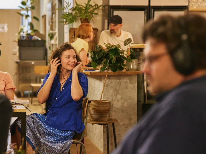 A woman in a blue dress listening to an audio piece with headphones, café ambiance in the background.