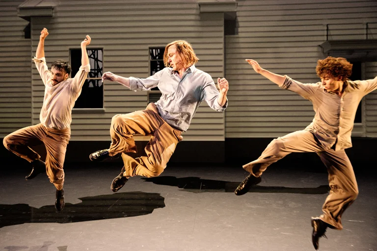 Three male performers — Marco Labellarte, Lars Eidinger, and Samuel Planas — leap in unison on a theater stage in front of a light-colored house façade. All are dressed in beige trousers and light shirts. Their powerful movement is captured mid-air, with raised arms and bent legs, frozen in a dynamic moment of choreography.