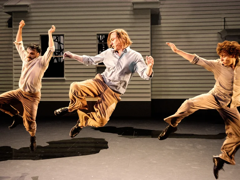 Drei männliche Performer – Marco Labellarte, Lars Eidinger und Samuel Planas – springen synchron auf einer Theaterbühne vor einer hellen Hausfassade. Alle tragen beigefarbene Hosen und helle Hemden. Ihre kraftvolle Bewegung ist in der Luft eingefroren, die Arme hoch erhoben und die Beine angewinkelt.