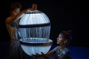 Szene aus der partizipativen Performance UNIVERS: Zwei Performerinnen öffnen ein großes, leuchtendes Objekt mit feinen, herabhängenden Fäden und laden zum stillen Beobachten ein.