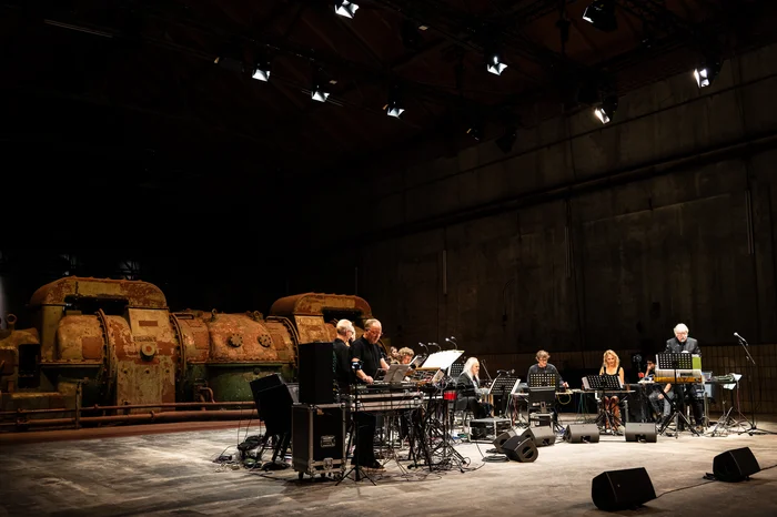 The Will Gregory Moog Ensemble performs on stage in a former industrial hall, illuminated by warm lighting. In the background, two large, rusty turbines are visible as the musicians operate electronic instruments like synthesizers and mixing consoles.